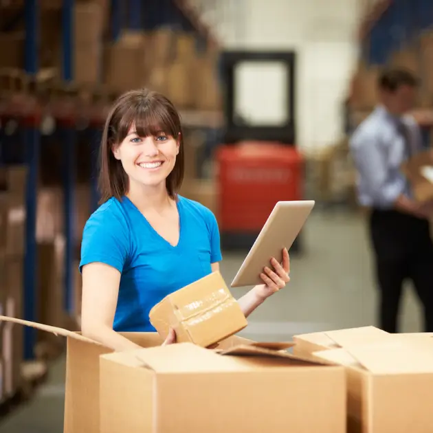A smiling woman in a blue shirt holding a tablet and a package, standing in a warehouse with boxes, representing efficient inventory management.